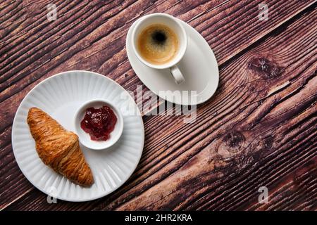 Un croissant, marmellata di fragole e caffè espresso su un piatto rotondo bianco su sfondo di legno preso dall'alto, una colazione preferita in tutto il mondo. Foto Stock