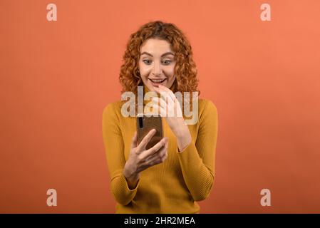 Una giovane e attraente ragazza caucasica rossa in un ponticello arancione utilizzando uno smartphone e la ragazza è scioccata isolato su uno sfondo arancione studio. Foto Stock