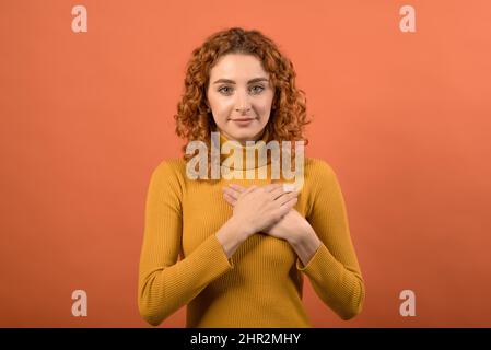 Giovane e attraente ragazza caucasica rossa in jumper arancione mostrando gesto di gratitudine mentre tenendo le mani al petto isolato sul backgrou studio arancione Foto Stock