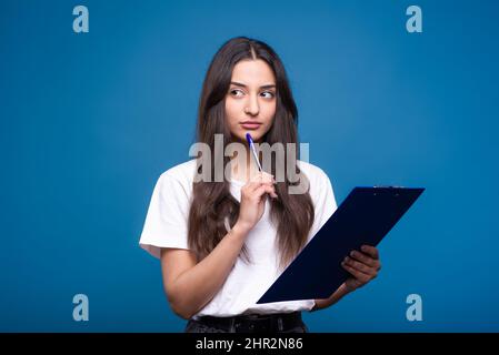 Bella ragazza bruna caucasica o araba in una t-shirt bianca tenendo una penna e un tablet nelle sue mani e pensando isolato su uno studio blu backgroun Foto Stock