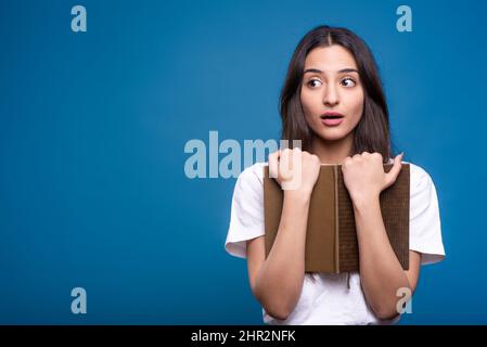 Attraente ragazza bruna caucasica o araba in t-shirt bianca tenendo libro e guardando verso lo spazio copia per la pubblicità isolato su blu studio backgr Foto Stock