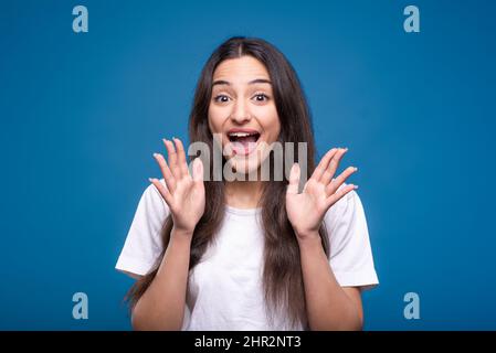 Attraente ragazza bruna caucasica o araba in una t-shirt bianca è scioccata e gioire nella vittoria isolato su uno sfondo blu studio. Foto Stock