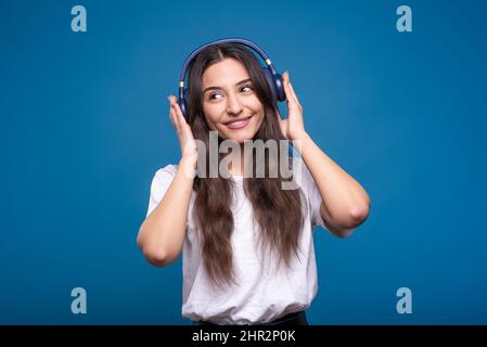 Bella ragazza bruna caucasica o araba in una t-shirt bianca che ascolta la sua musica preferita con le cuffie e sorride isolato su uno studio blu retro Foto Stock