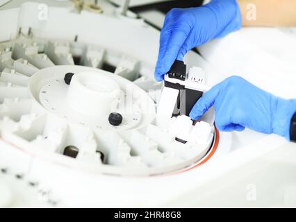 l'assistente di laboratorio in guanti di gomma blu aggiunge un reagente alla centrifuga di un moderno analizzatore biochimico. Foto Stock