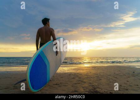 Uomo asiatico che tiene un sup board e paddle e camminare sulla spiaggia. Sullo sfondo, l'oceano e il tramonto. Vista posteriore. Surf estivo. Foto Stock