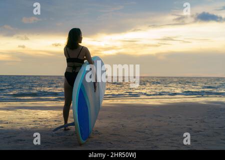 Donna asiatica che tiene una tavola di sup e paddle e cammina sulla spiaggia. Sullo sfondo, l'oceano e il tramonto. Vista posteriore. Surf estivo. Foto Stock