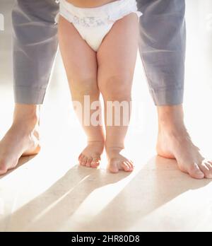 Primi passi. Immagine ritagliata delle gambe di un bambino che cammina con un genitore che sostiene da dietro. Foto Stock