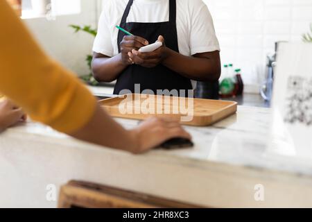 Metà della sezione del barista afro-americano che prende ordine dal cliente al banco in caffetteria Foto Stock