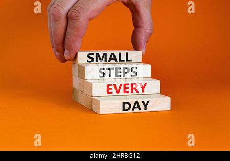 Simbolo piccoli passi ogni giorno. Parole di concetto piccoli passi ogni giorno su blocchi di legno. Mano dell'uomo d'affari. Bella tabella arancione sfondo arancione. Piccola Foto Stock