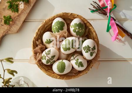 Uova di Pasqua crude con erbe attaccate a loro in un cestino, pronto per essere tinto con bucce di cipolla, vista dall'alto Foto Stock