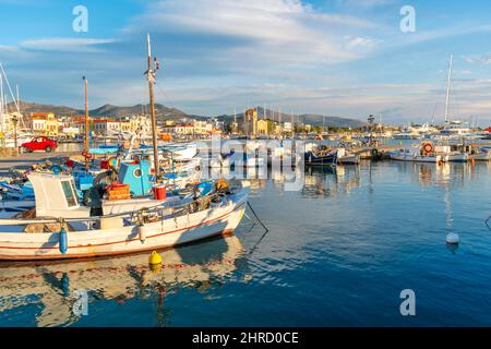 Colorate barche da pesca rosse, bianche e blu fiancano il porto dell'isola greca di Egina, in Grecia, con il piccolo villaggio storico alle spalle Foto Stock