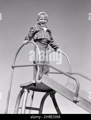 1960S RAGAZZO BIONDO SORRIDENTE CHE INDOSSA UNA GIACCA A PLAID IN PIEDI IN CIMA ALLA TAVOLA SCORREVOLE NEL PARCO GIOCHI GUARDANDO LA MACCHINA FOTOGRAFICA - J9703 HAR001 HARS SLIDE SANO FELICE GIOIA LIFESTYLE SODDISFAZIONE ORGOGLIOSA COPIA SPAZIO FULL-LENGTH MASCHI NERVOSA PLAID FIDUCIA B&W OCCHIO CONTATTO ATTIVITÀ FELICITÀ FISICA ALLEGRO AVVENTURA FORZA CORAGGIO ECCITAZIONE BASSO ANGOLO RICREAZIONE SCORRIMENTO ORGOGLIO SORRISI FLESSIBILITÀ GIOIOSO MUSCOLI CRESCITA GIOVANI NERO E. BIANCO CORAGGIOSO ETNIA CAUCASICA HAR001 VECCHIO STILE Foto Stock