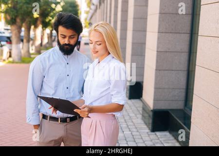 Ritratto verticale medio del ritratto attraente giovane donna d'affari bionda che spiega qualcosa sulla clipboard al compagno maschio bello di affari Foto Stock