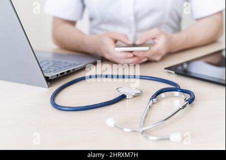 Una donna irriconoscibile si siede su una scrivania e utilizza uno smartphone. Primo piano delle mani di un medico con un telefono. Stetoscopio tablet e notebook Foto Stock