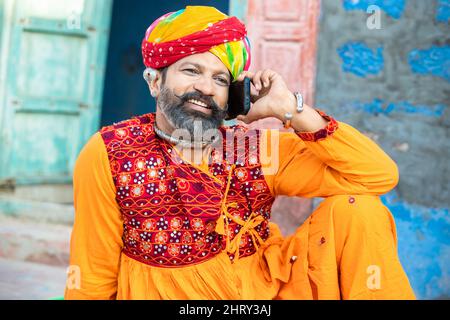 Felice uomo maturo indiano del nord indossando abiti colorati e turbante parlare su smartphone, sorridente tradizionale maschio chiamata su android smart mobile pho Foto Stock