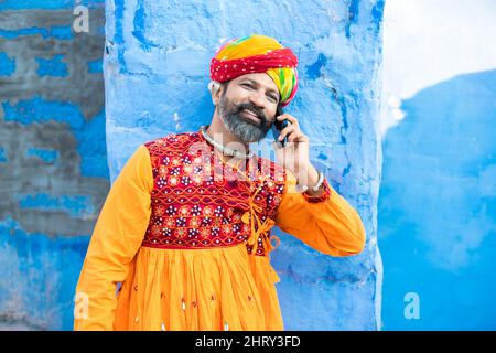 Felice uomo maturo indiano del nord indossando abiti colorati e turbante parlare su smartphone, sorridente tradizionale maschio chiamata su android smart mobile pho Foto Stock