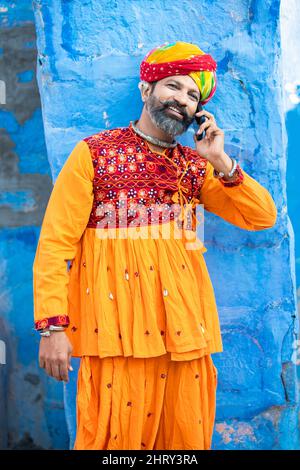 Felice uomo maturo indiano del nord indossando abiti colorati e turbante parlare su smartphone, sorridente tradizionale maschio chiamata su android smart mobile pho Foto Stock