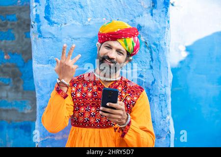 Felice uomo maturo indiano tradizionale indossando abiti colorati e turbante utilizzando smartphone, sorridente maschio tenendo android smartphone mentre in piedi Foto Stock