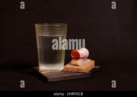 Bicchiere sfaccettato di vodka, panino con pane di segale, aringhe, prosciutto Foto Stock