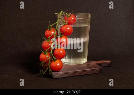 Un bicchiere sfaccettato di vodka e un rametto di pomodori ciliegini su un supporto di legno Foto Stock