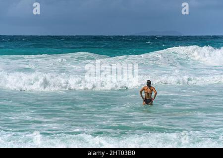 Giovane donna con costume da bagno nero, in piedi in mare turchese con grandi onde Foto Stock
