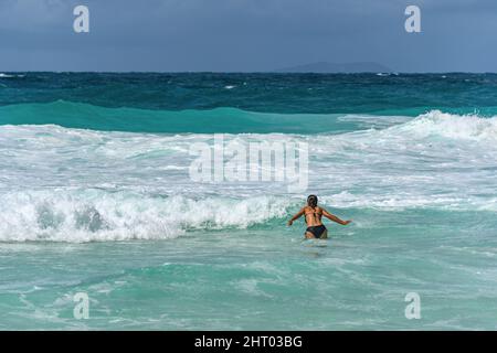 Giovane donna con costume da bagno nero, in piedi in mare turchese con grandi onde Foto Stock