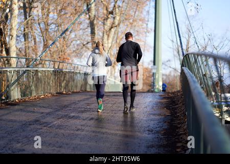Stoccarda, Germania - 06 gennaio 2022: Giovane coppia che fa sport insieme nel parco. Uomo e donna che jogging su un ponte alla luce del giorno. Vista posteriore. STU Foto Stock
