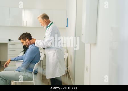 Vista laterale di adulto maturo maschio terapista manuale girando e piegando la testa di paziente maschio per controllare il suo collo ferito. Foto Stock