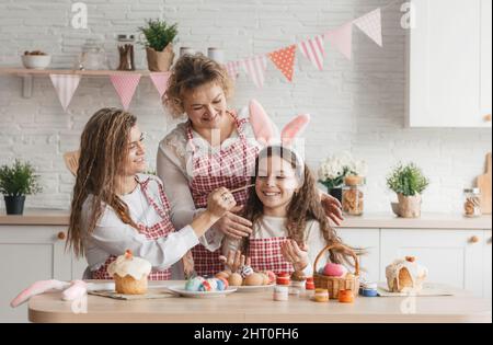 Una mamma felice con le sue figlie adulte dipinge le uova di Pasqua a casa in cucina. Foto Stock