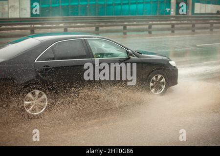 L'auto passa attraverso la pozzanghera. Spruzzi da sotto le ruote. Pioggia in autostrada. Strada sporca. Alta velocità in pista. Foto Stock