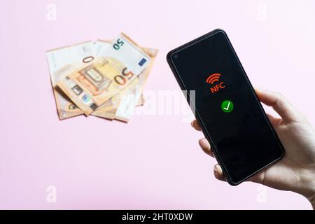 La ragazza tiene in mano un cellulare con l'icona NFC sullo schermo e alcune banconote in euro sullo sfondo Foto Stock