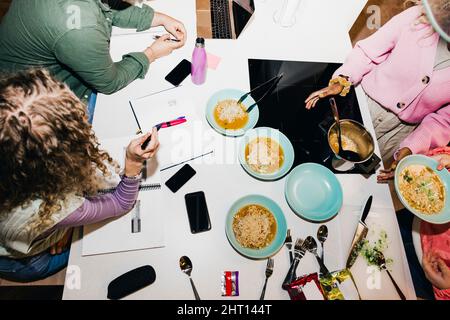 Vista ad angolo alto di giovani amici multirazziale che hanno tagliatelle mentre studiano al tavolo da pranzo in dormitorio college Foto Stock