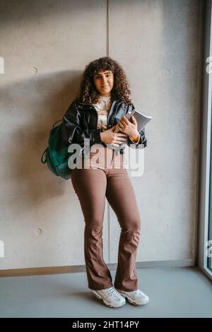 Ritratto a tutta lunghezza della giovane donna che tiene un libro e uno zaino appoggiato su una parete grigia Foto Stock
