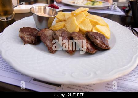 Bistecca di controfiletto alla griglia con patate a fette arrostite, il tutto su un piatto bianco. Una ciotola con salsa calda. Cucina spagnola. Foto Stock