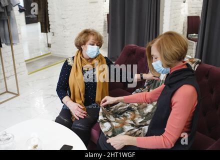 Due donne anziane in maschere mediche stanno guardando i vestiti seduti su un divano in un negozio di abbigliamento. Shopping e shopping Foto Stock