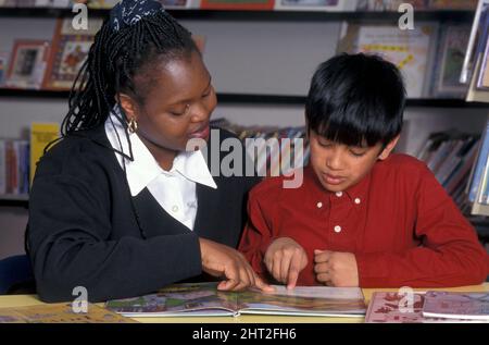 Donna africana che legge con ragazzo asiatico in biblioteca che ha esigenze speciali Foto Stock