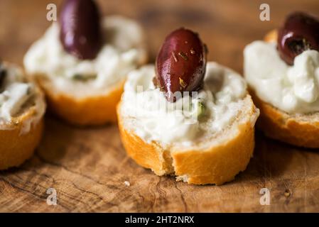 Piccolo pezzo di pane con tzatziki spalmato e frutta di oliva su tavola di legno da cucina di olivo, cucina greca tradizionale. Primo piano. Foto Stock