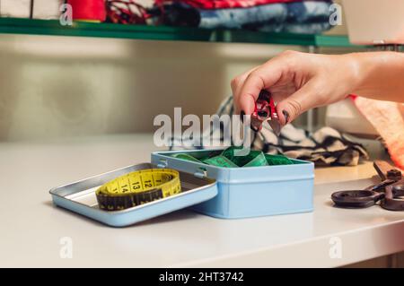 primo piano di mani caucasiche femmina tenendo forbici che sta prendendo da una cassetta degli attrezzi tessile sul tavolo in un laboratorio di cucito, spazio copia. Foto Stock