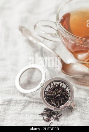 Tazza di vetro con tè nero caldo e infusore con tè sciolto su asciugamano bianco da cucina Foto Stock