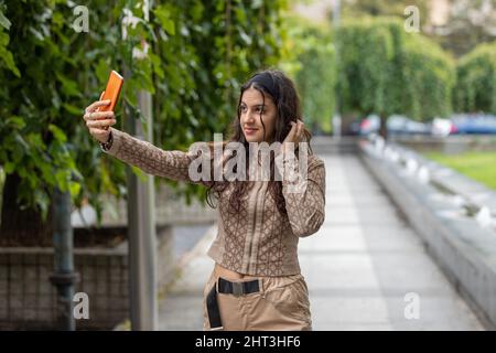 Ragazza sorridente che guarda lo smartphone, toccando e pettinando i capelli usando il cellulare come uno specchio per la strada Foto Stock