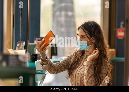 Cute teen indossare maschera medica seduta in caffè toccare e pettinare i capelli utilizzando smartphone come specchio Foto Stock