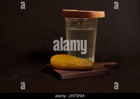 Un bicchiere sfaccettato di vodka, pane di segale e cetriolo sottaceto su un piedistallo di legno Foto Stock