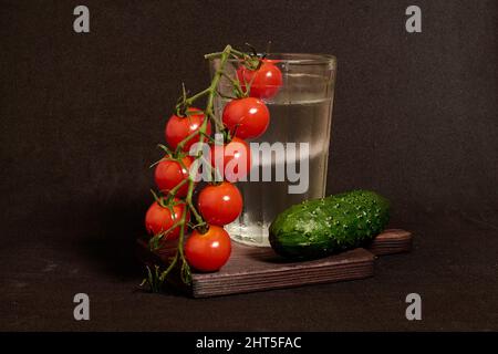 Un bicchiere sfaccettato di vodka, un ramo di pomodori ciliegini e un cetriolo fresco Foto Stock