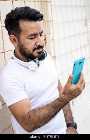 Uomo latino con una barba, guardando il suo cellulare appoggiato a un muro Foto Stock