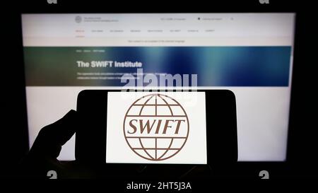 Persona che detiene il cellulare con il logo del sistema di transazioni bancarie SWIFT sullo schermo di fronte alla pagina web. Mettere a fuoco sul display del telefono. Foto Stock