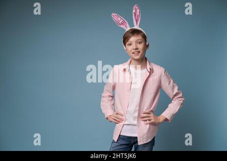 Foto giovane ragazzo piacevole con divertente umore infantile gesturing con le mani e guardando la macchina fotografica, con le orecchie rosa di un coniglietto pasquale con un bendaggio sulle orecchie in una camicia rosa casual isolato su blu Foto Stock