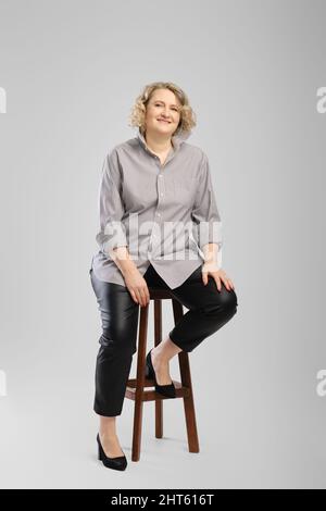 Donna anziana in camicia grigia e pantaloni in pelle sintetica si siede su un'alta sedia di legno su sfondo grigio studio Foto Stock