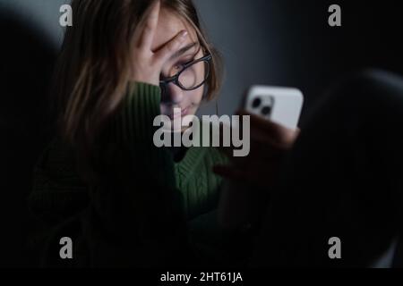 Ragazza piccola triste, sola al buio, seduta e con smartphone. Foto Stock