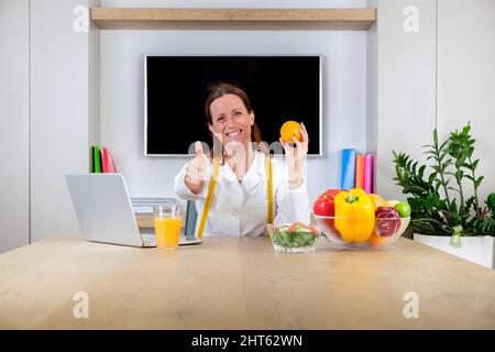 Ritratto di nutrizionista sorridente che tiene un arancione. Femmina dietista sorridente mostrando frutta e facendo pollice su e segno di approvazione con le dita Foto Stock