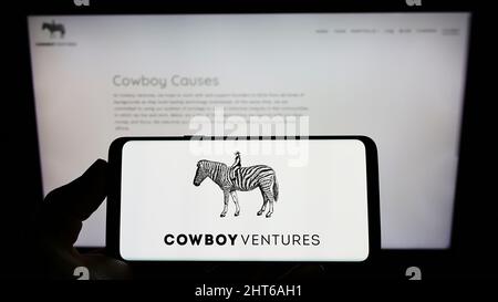 Persona che tiene il cellulare con il logo della società americana di capitali di rischio Cowboy Ventures sullo schermo di fronte al sito web. Mettere a fuoco sul display del telefono. Foto Stock